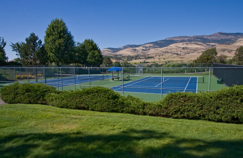 A scenic view of outdoor tennis courts surrounded by greenery and mountains under a clear blue sky.