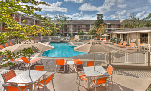 A sunny pool area with orange lounge chairs and umbrellas, surrounded by a hotel and trees under a blue sky.