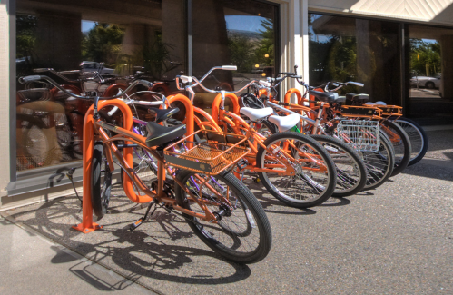 A row of bicycles parked at an orange bike rack outside a building, with sunlight reflecting off the windows.