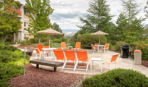 Outdoor seating area with orange lounge chairs, tables, and umbrellas surrounded by greenery and gravel pathways.