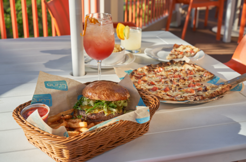 A burger with fries in a basket, a cocktail, and a pizza on a table with orange chairs in the background.