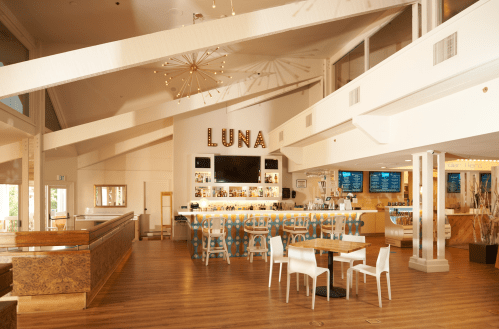 Bright, modern restaurant interior with a bar, tables, and a large "LUNA" sign on the wall. Warm wooden tones and decor.