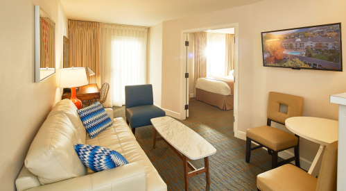 A cozy hotel suite featuring a living area with a sofa, coffee table, and a TV, plus a bedroom visible in the background.