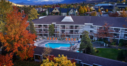Aerial view of a hotel with a pool, surrounded by colorful autumn trees and mountains in the background.