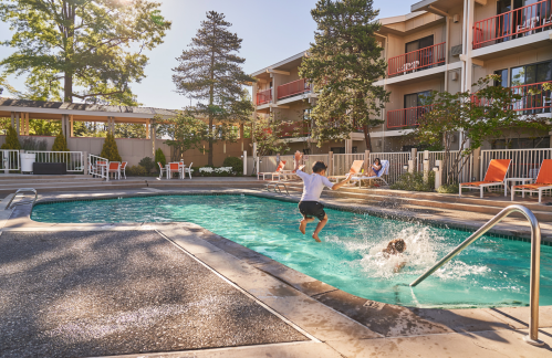 A child jumps into a pool while a dog splashes nearby, with lounge chairs and a hotel in the background.