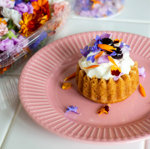 A small cake topped with whipped cream and colorful edible flowers on a pink plate, surrounded by more flowers.