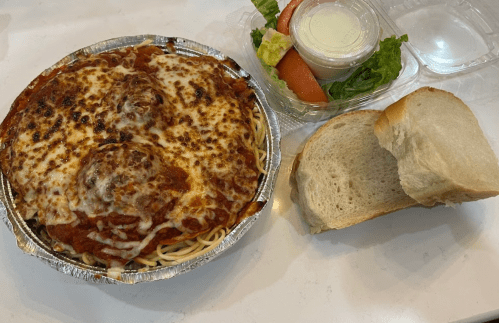 A foil container with spaghetti and meatballs topped with cheese, alongside a salad and a slice of bread.