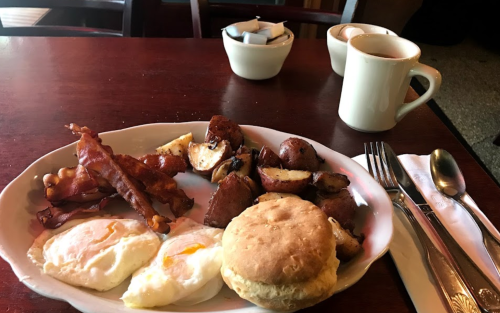 A plate of breakfast with two poached eggs, bacon, roasted potatoes, and a biscuit, alongside a cup of coffee.
