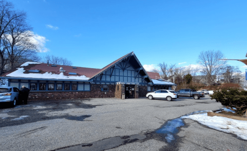 A large, brick building with a sloped roof, surrounded by a parking lot and trees, under a clear blue sky.