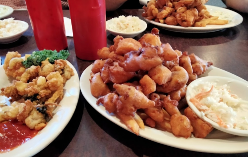 A table with plates of fried seafood, fries, coleslaw, and condiments in red bottles.