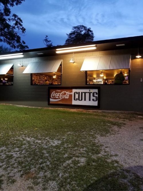 A dimly lit building with a Coca-Cola sign and "CUTTS" displayed, surrounded by grass and trees at dusk.