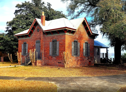 Historic brick building with a gabled roof, surrounded by trees and a grassy area, featuring large windows and a porch.