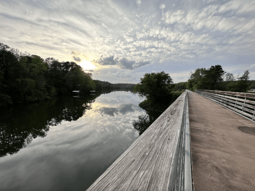 A serene river scene at sunset, with clouds reflecting on the water and a wooden walkway along the bank.