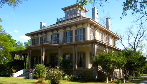 A large, historic house with a multi-tiered roof, surrounded by greenery and a clear blue sky.