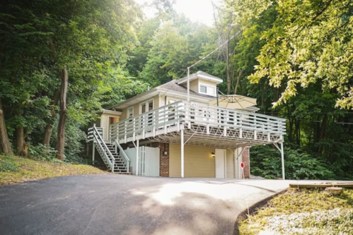 A modern house with a balcony, surrounded by lush green trees, set on a sloped driveway.