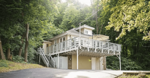 A modern house with a balcony, surrounded by lush green trees, set on a sloped driveway.