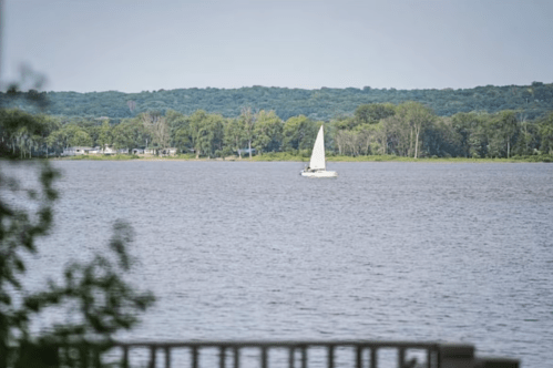 A small sailboat glides across a calm lake, surrounded by lush green trees in the background.