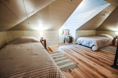 Cozy attic bedroom with two beds, wooden floors, and soft lighting, featuring sloped ceilings and decorative rugs.