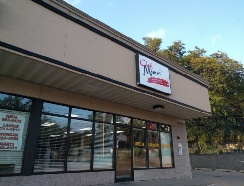 Exterior of a restaurant named "Club Menussa," featuring large windows and a sign, with trees in the background.