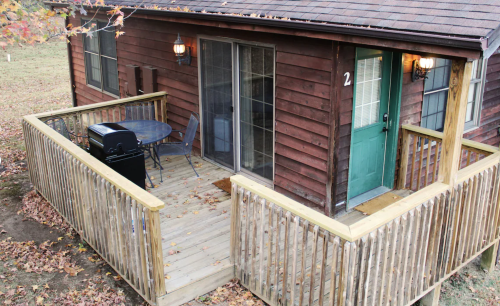 A wooden cabin with a green door, deck, and outdoor seating area, surrounded by autumn foliage.