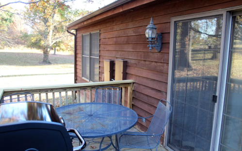 A wooden deck with a grill, a round table, and chairs, surrounded by trees and a grassy area.
