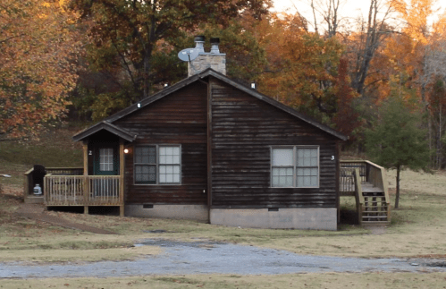 A rustic wooden cabin surrounded by autumn trees, featuring a porch and a stone chimney.