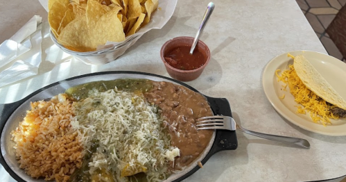 A plate of enchiladas with rice and beans, a side of salsa, and a basket of tortilla chips on a table.