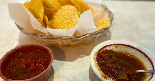 A basket of tortilla chips next to two bowls of salsa, one red and one brown, on a table.