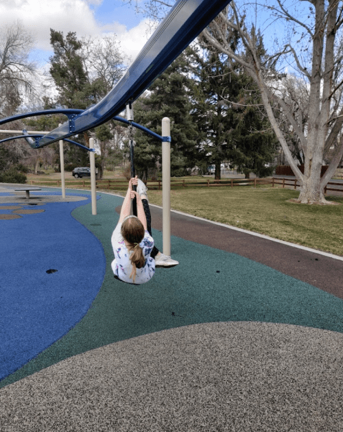 A child swings on a playground swing, with trees and a grassy area in the background under a partly cloudy sky.