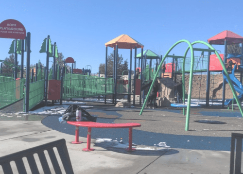 A colorful playground with slides and climbing structures, surrounded by green fencing and a clear blue sky.