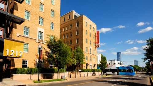 A city street featuring brick buildings, trees, and a blue bus, with a skyline in the background under a clear sky.