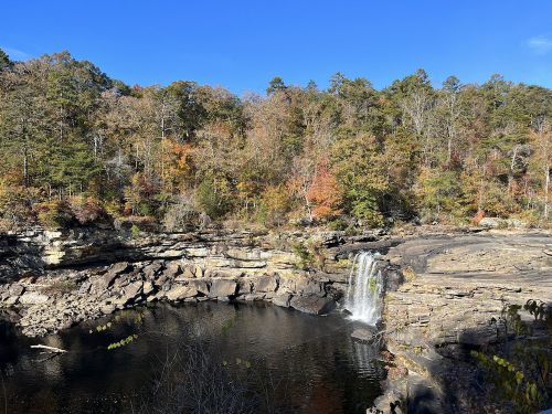 A serene waterfall cascades into a calm pool, surrounded by autumn foliage and rocky terrain under a clear blue sky.
