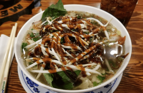 A bowl of pho topped with herbs, bean sprouts, and drizzled with sauce, alongside chopsticks and a drink.