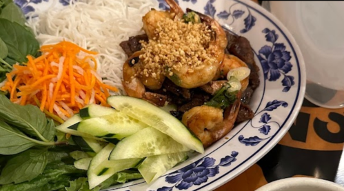 A colorful plate featuring shrimp, grilled meat, rice noodles, fresh herbs, cucumber, and shredded carrots.