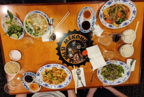 A table set with various Asian dishes, including noodles, stir-fried vegetables, and sauces, surrounded by drinks.