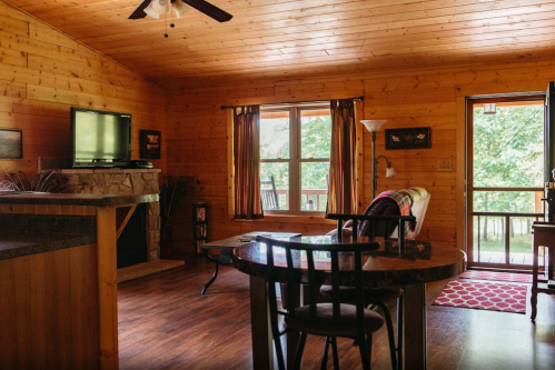 Cozy wooden cabin interior with a dining table, TV, and door leading to a scenic outdoor view.