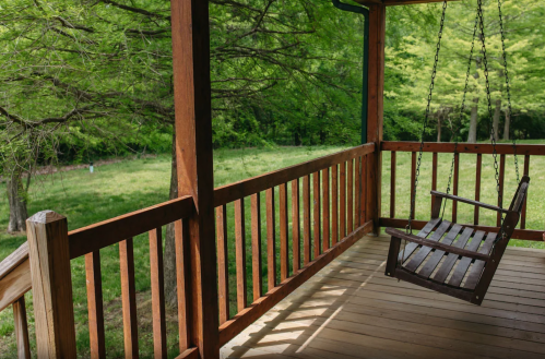A wooden porch swing hangs on a rustic porch, overlooking a lush green field and trees in the background.