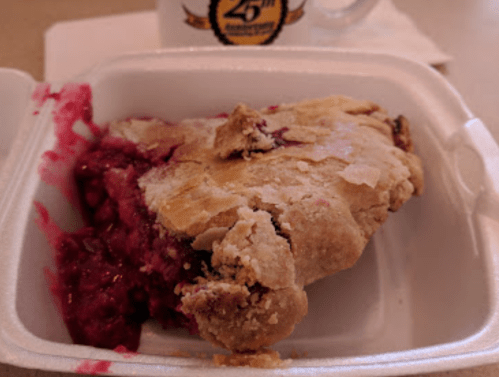 A slice of pie with a flaky crust and berry filling, served in a white takeout container.