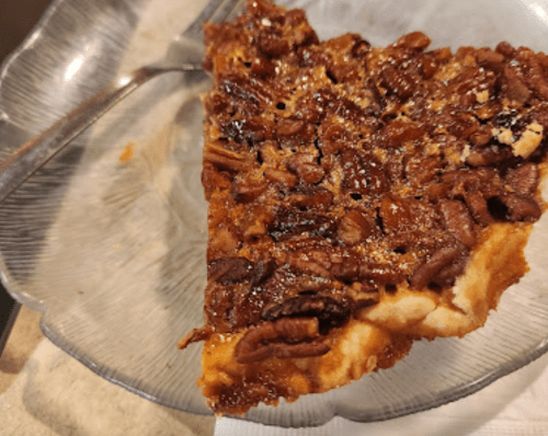A slice of pecan pie on a clear glass plate, with a fork resting beside it.