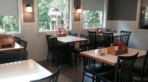 A cozy restaurant interior with empty tables, black chairs, and large windows overlooking greenery.