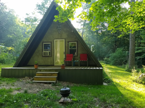 A cozy A-frame cabin surrounded by trees, featuring a porch with red chairs and a small grill in the foreground.