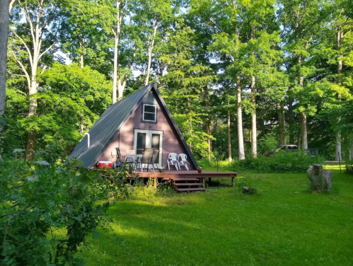 A cozy A-frame cabin surrounded by lush green trees and a grassy yard, featuring a deck with chairs.