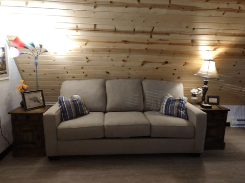 A cozy attic living space featuring a beige sofa with plaid pillows, a lamp, and wooden side tables against a wooden wall.