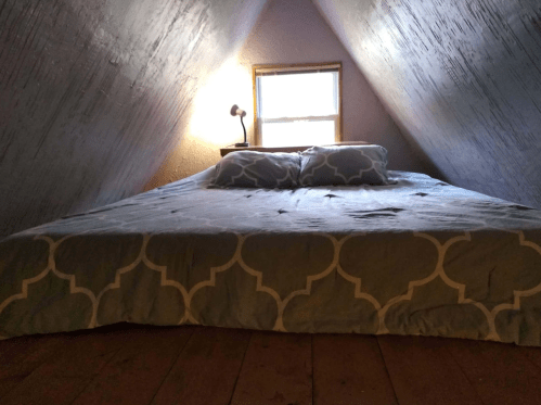 Cozy attic bedroom with a sloped ceiling, a bed with a patterned blanket, two pillows, and a lamp by the window.