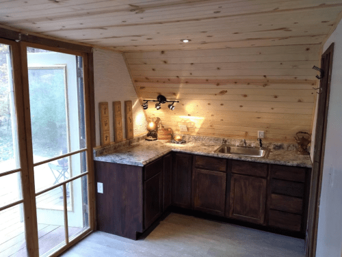 Cozy kitchen with wooden walls, granite countertops, and a sink, featuring a large window and sliding glass door.