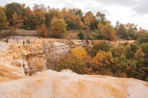 A rocky cliff with people exploring, surrounded by trees displaying autumn colors under a cloudy sky.