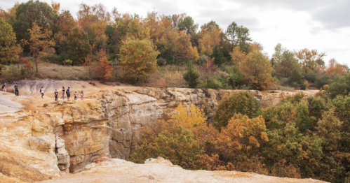 A rocky cliff with people exploring, surrounded by trees displaying autumn colors under a cloudy sky.