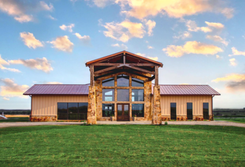 A modern building with a wooden frame and stone accents, set against a blue sky with clouds. Green grass in the foreground.