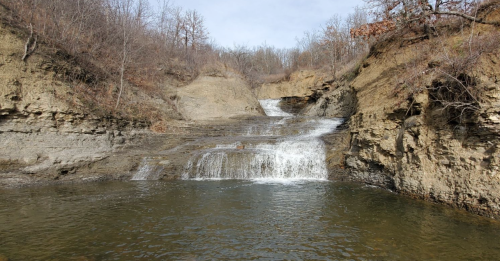 A serene waterfall cascading over rocky terrain into a calm pool, surrounded by bare trees and earthy cliffs.