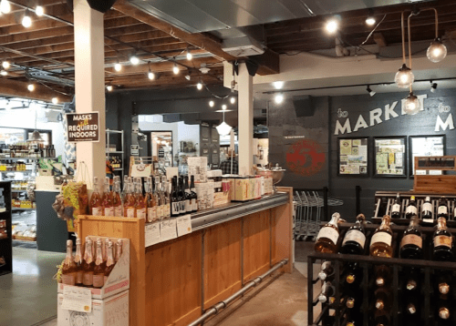 A cozy market interior featuring a wooden counter, shelves of wine, and a sign indicating mask requirements.